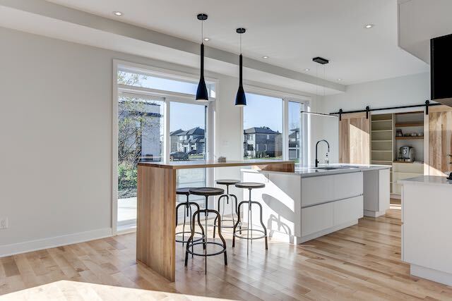 Cuisine moderne avec parquet clair, îlot blanc et tabourets de bar. Grandes fenêtres et suspensions.