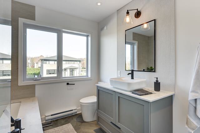 Salle de bain moderne avec vanité grise, lavabo blanc, miroir noir, fenêtre et mur carrelé blanc.