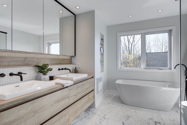 Salle de bain moderne avec vanité en bois clair, lavabos blancs et baignoire autoportante près d'une fenêtre.