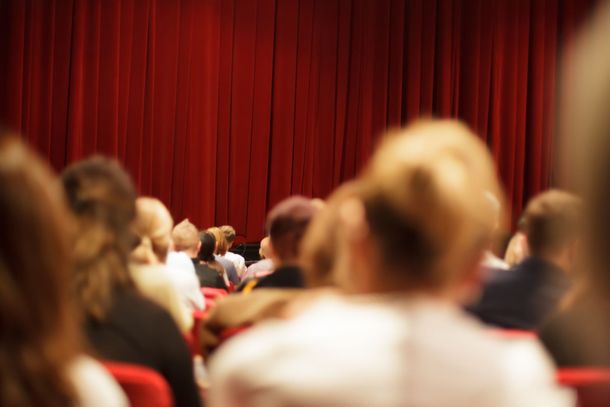 Audience seated before a red theater curtain, viewed from the back of the hall Audience seated before a red theater curtain, viewed from the back of the hall