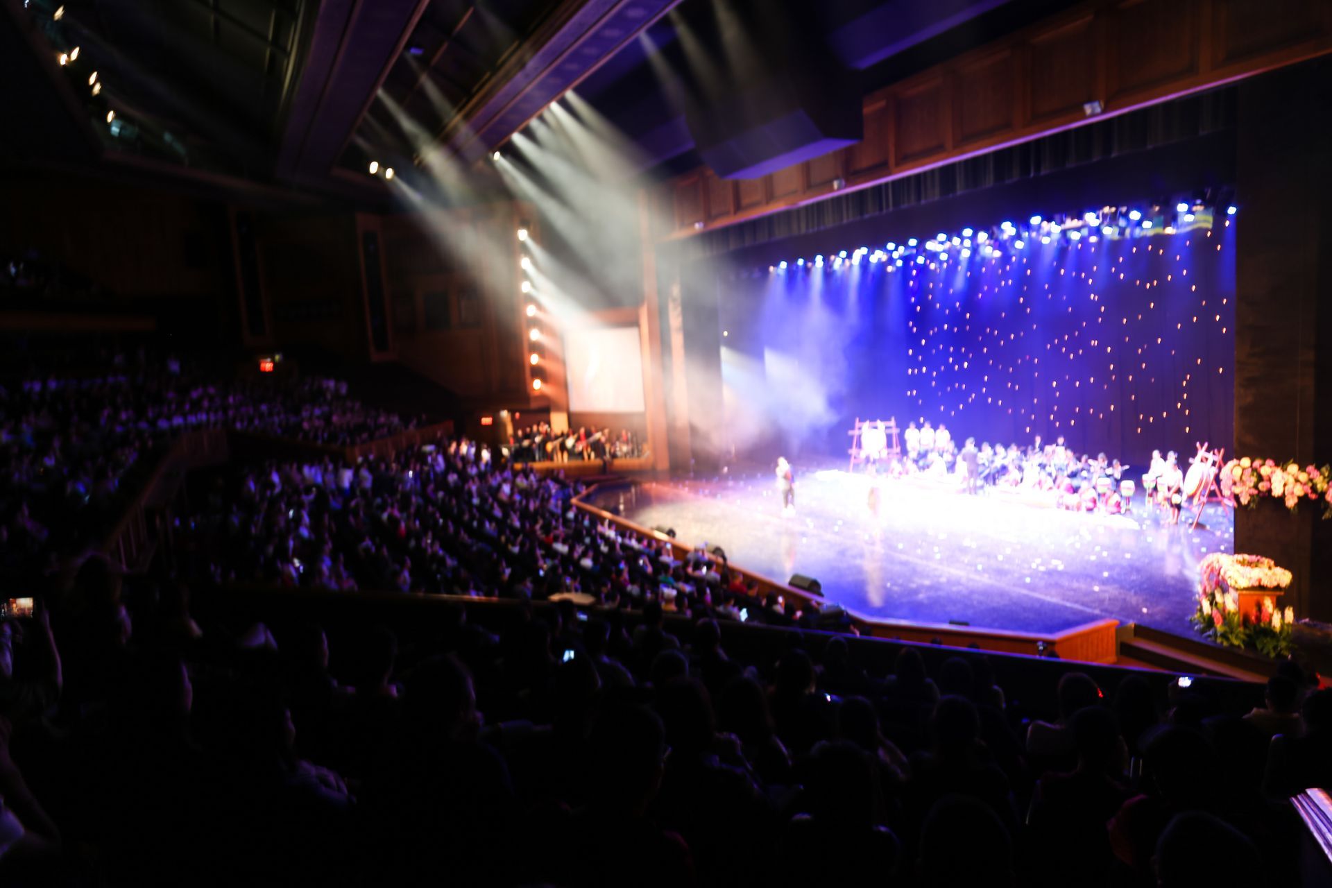 Auditorium with a brightly lit stage, blue backdrop, and a large audience watching a performance