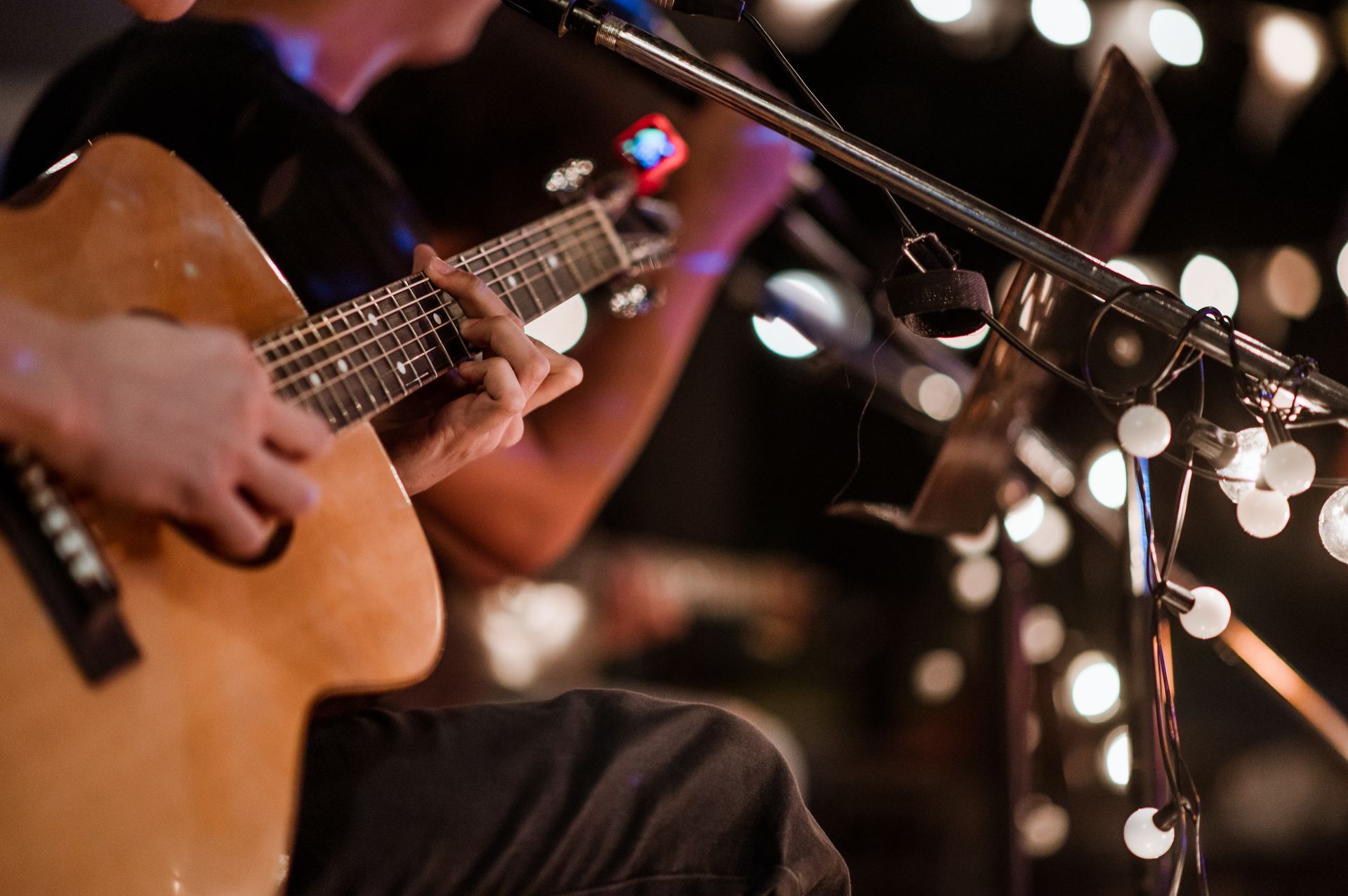 Musician playing acoustic guitar beside warm string lights at a dimly lit performance