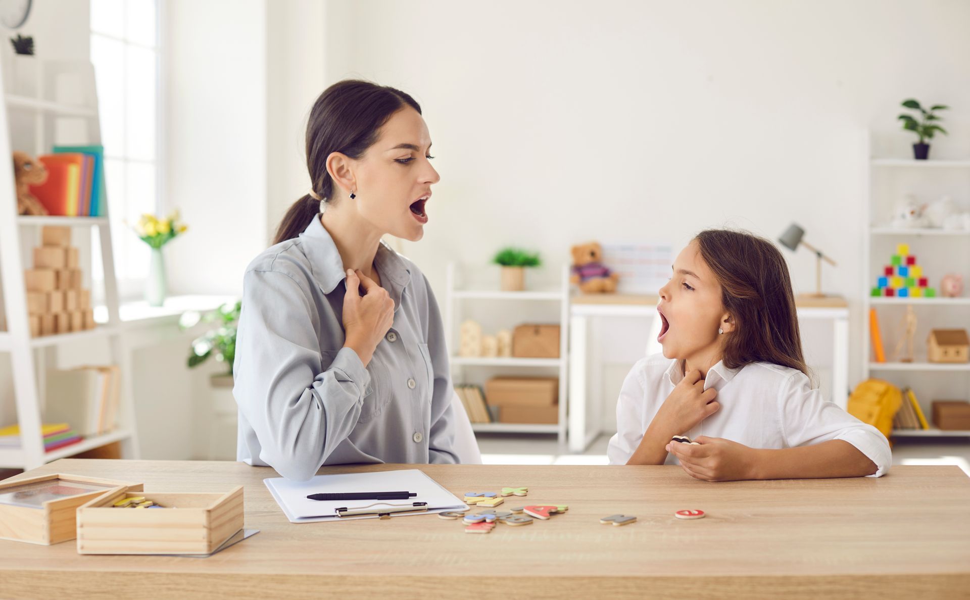 Teacher and child in a bright classroom, talking at a table with books and pencils.