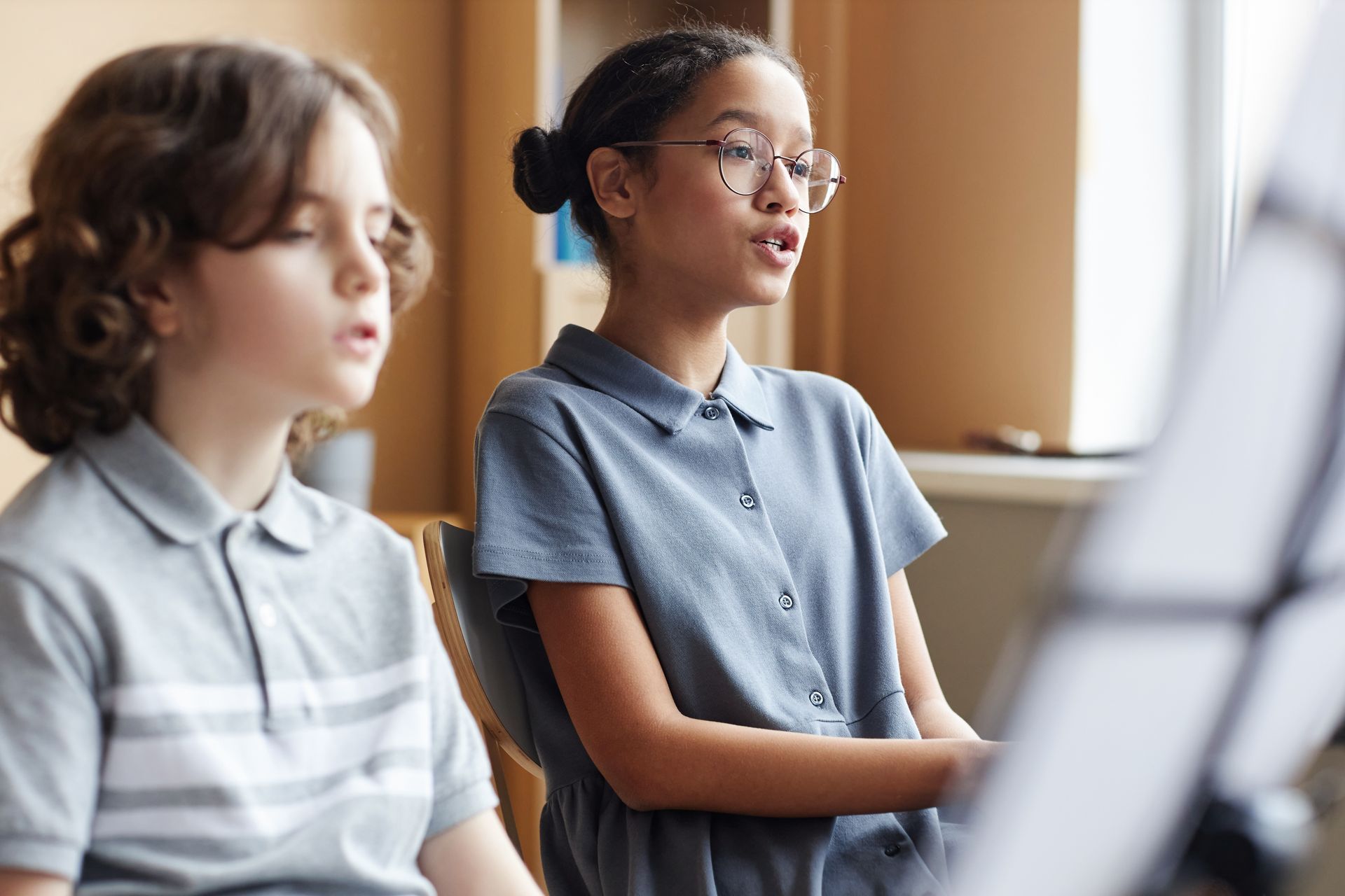 Two children in gray shirts seated in a classroom, one speaking while music stands blur in the foreground