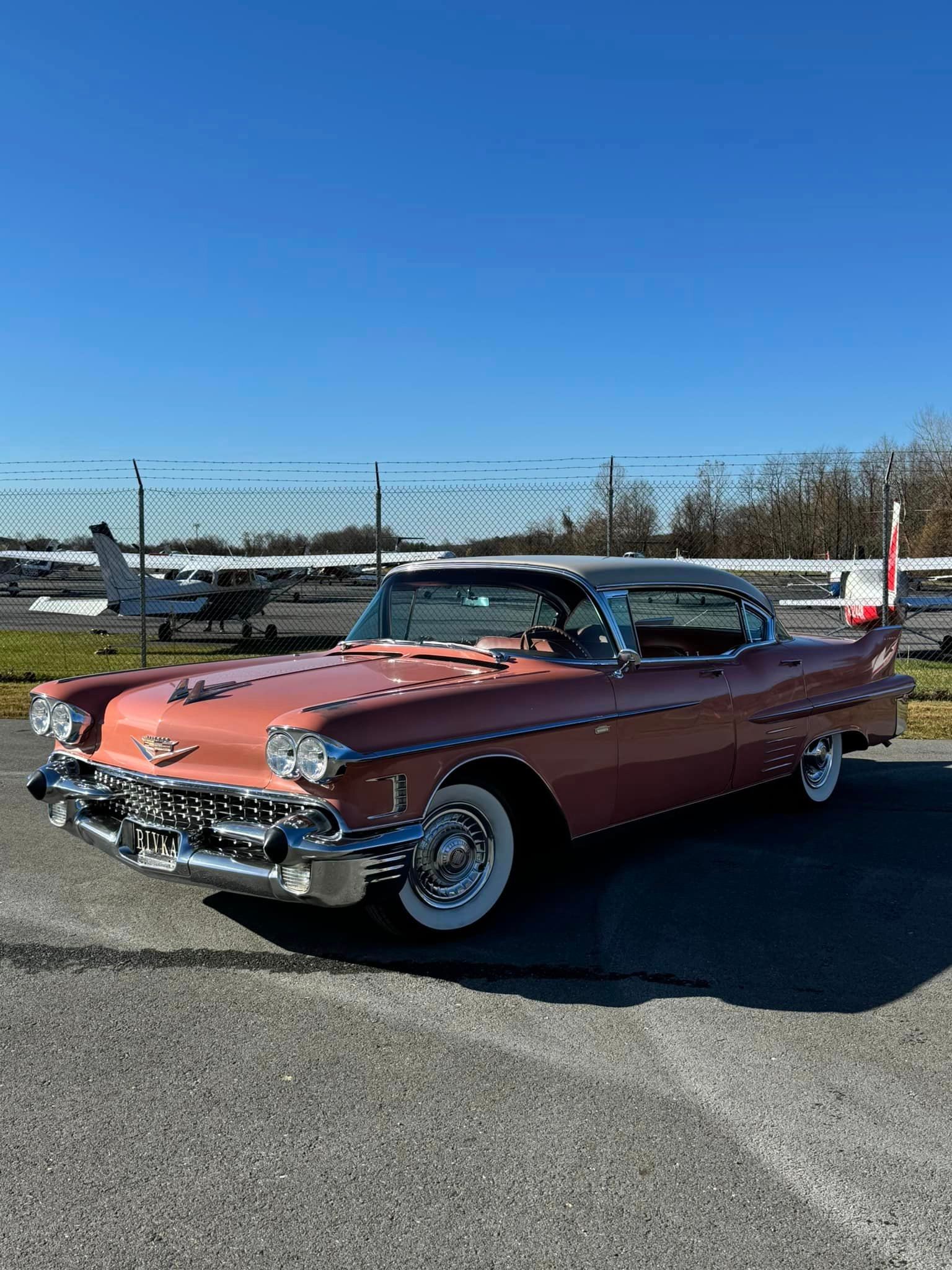A pink cadillac is parked in a parking lot next to a plane.