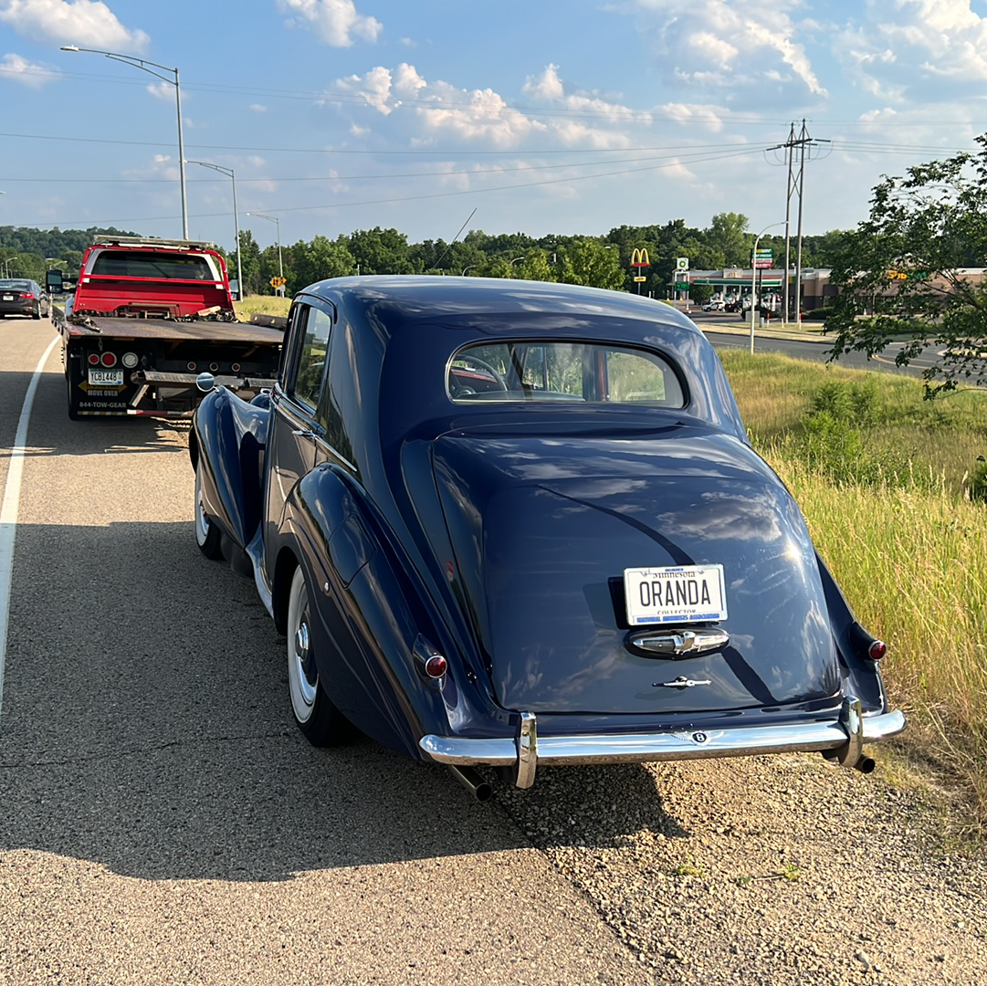 Blue classic car on a roadside tow truck with a red truck ahead under a partly cloudy sky