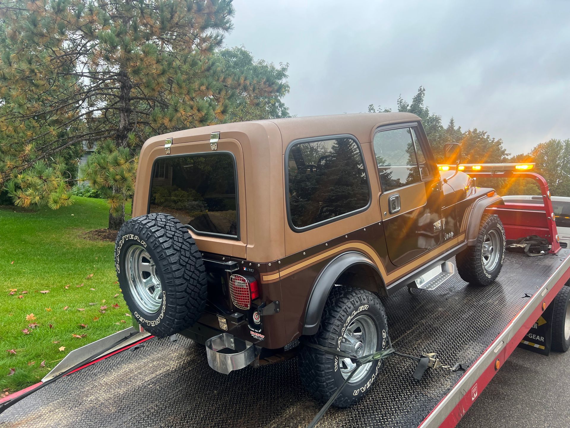 Brown Jeep Wrangler on a tow truck in a rainy, wooded roadside setting