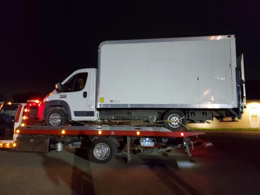White box truck on a tow truck at night, lit by red lights on a dark road.