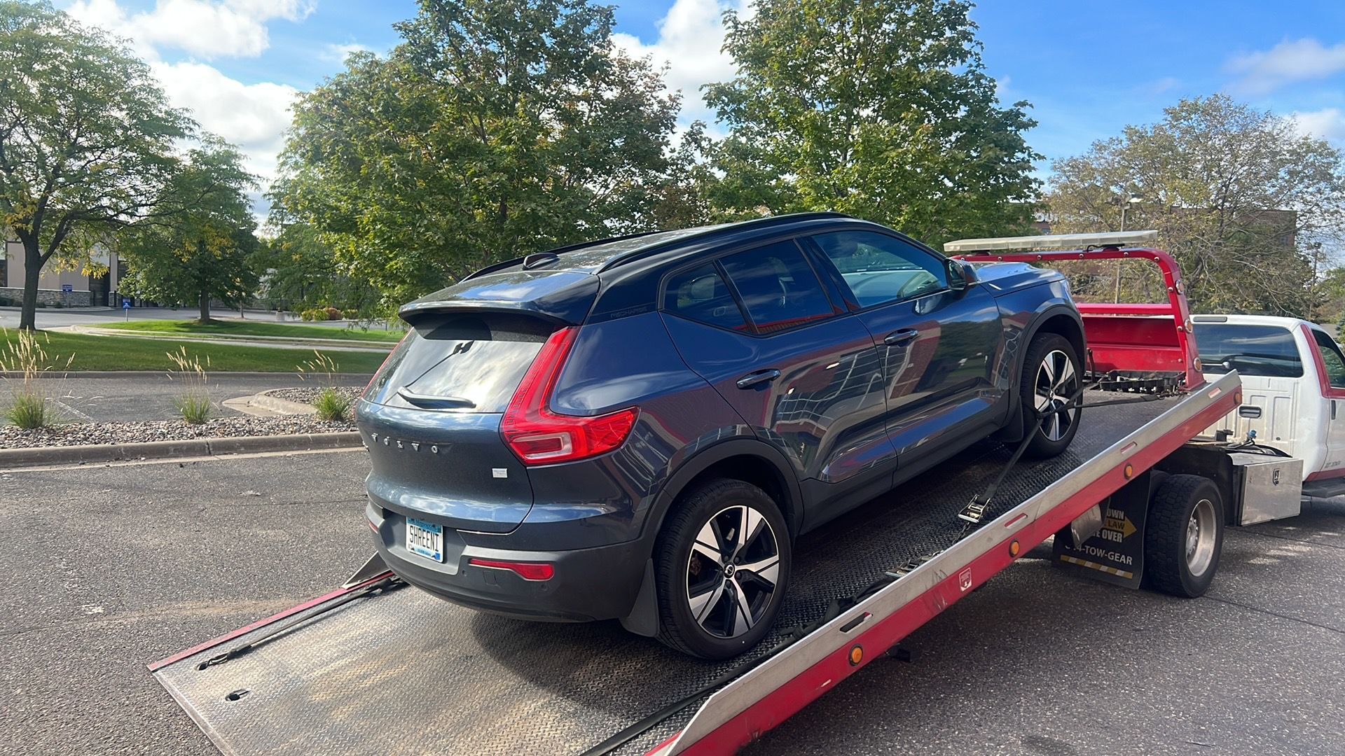 Dark blue SUV on a flatbed tow truck in a parking lot, with trees and houses in the background