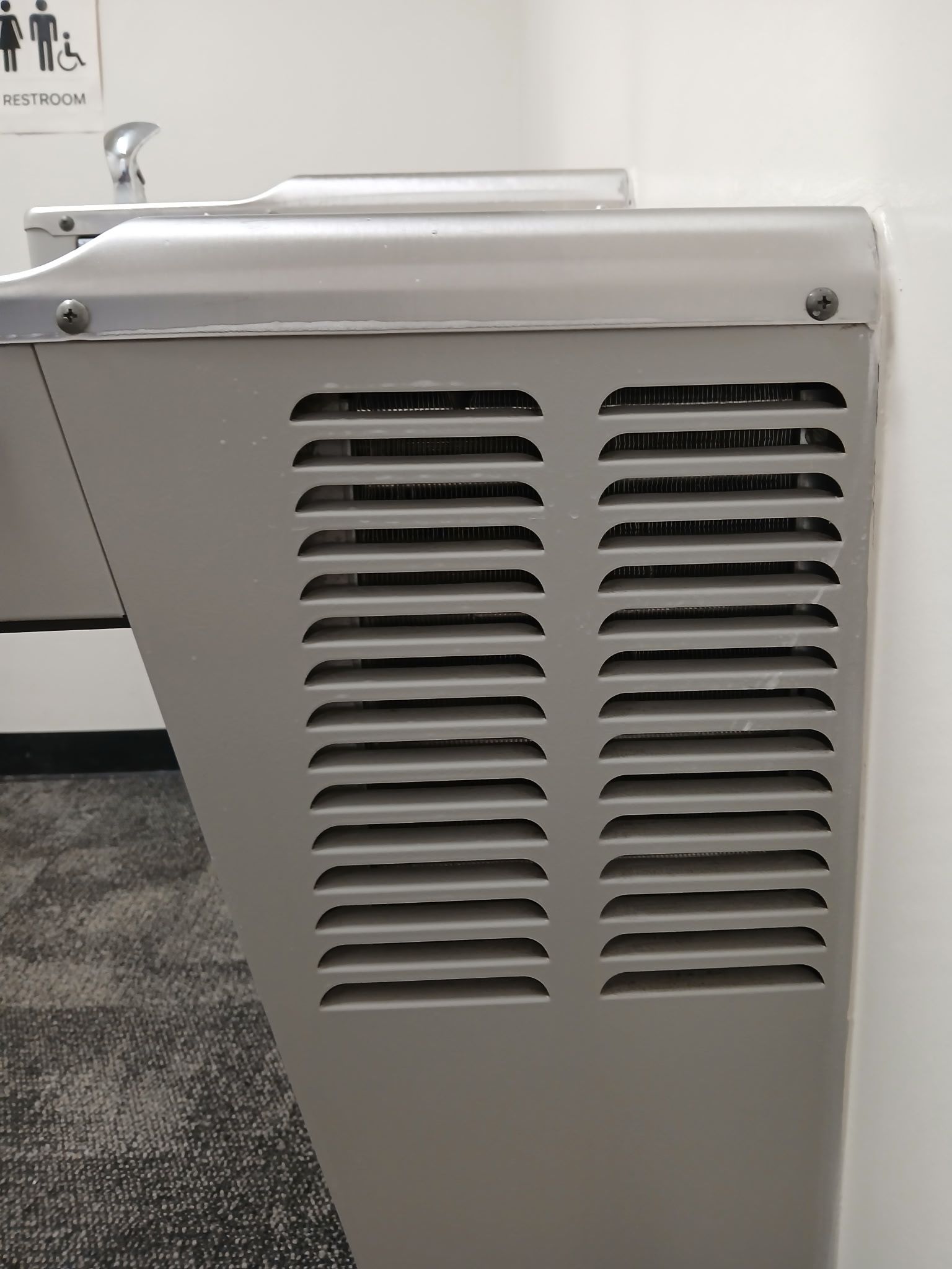 A light gray metal water fountain with side air vents mounted against a white wall.
