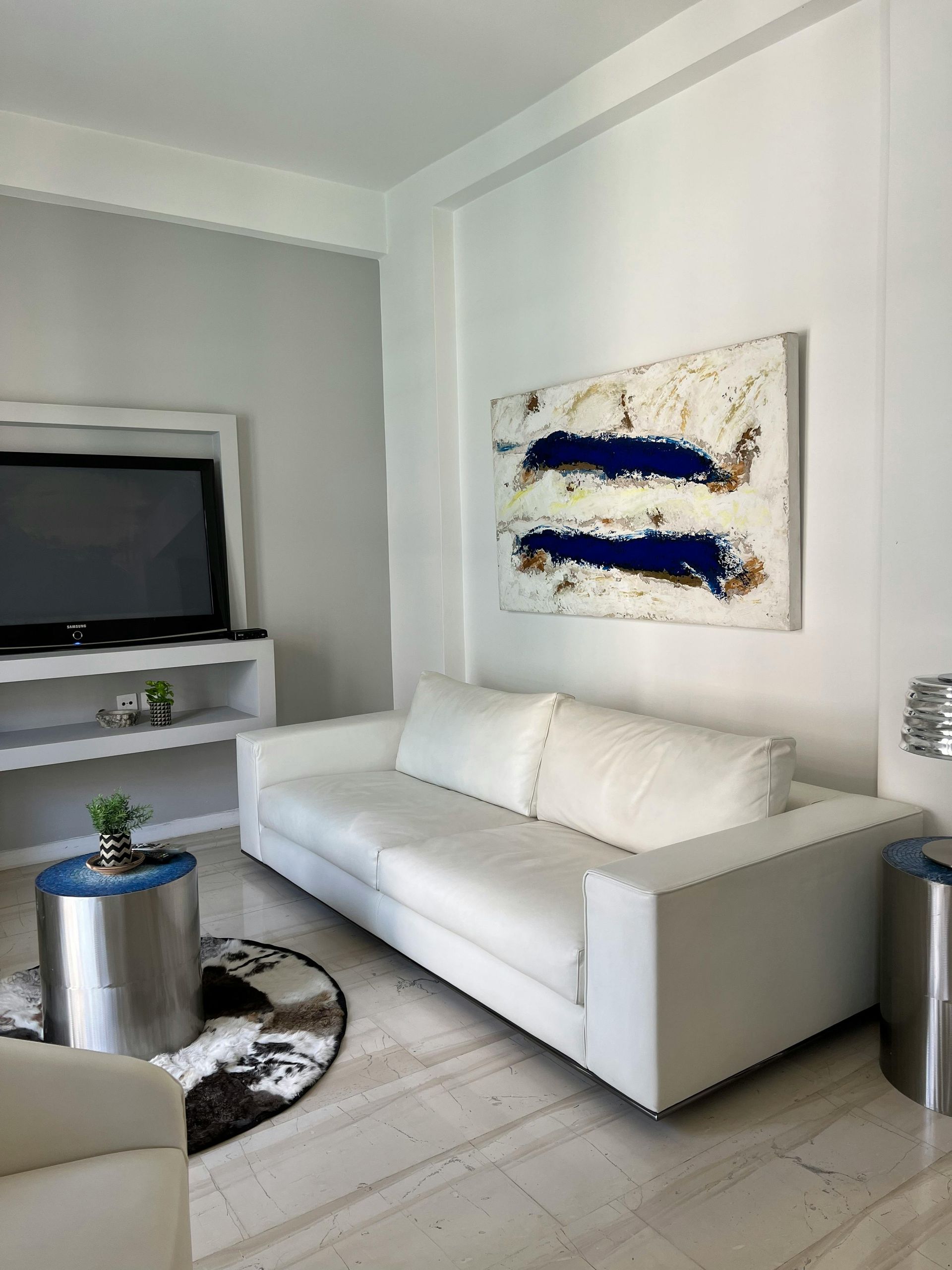 A modern living room with a white sofa, abstract blue and white art, a TV on a white shelf, and a round metallic table.