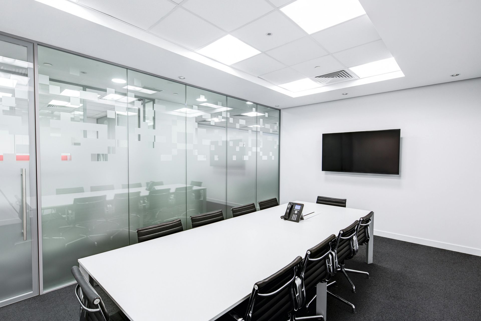 A modern, minimalist meeting room with a long white table, black office chairs, glass walls, and a wall-mounted TV.