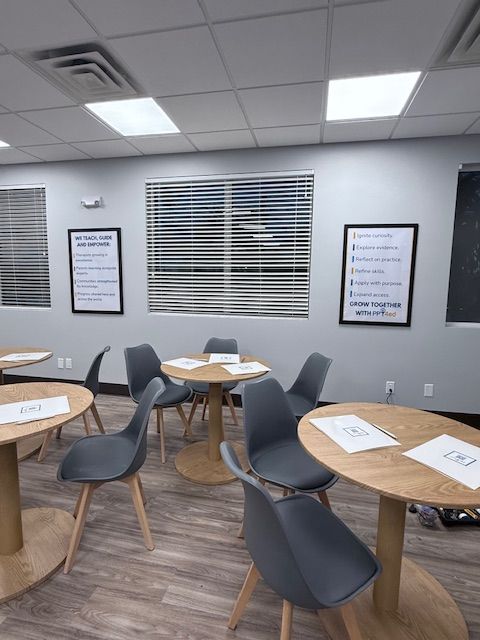 Room with round tables, gray chairs, blinds, posters, and overhead lighting.