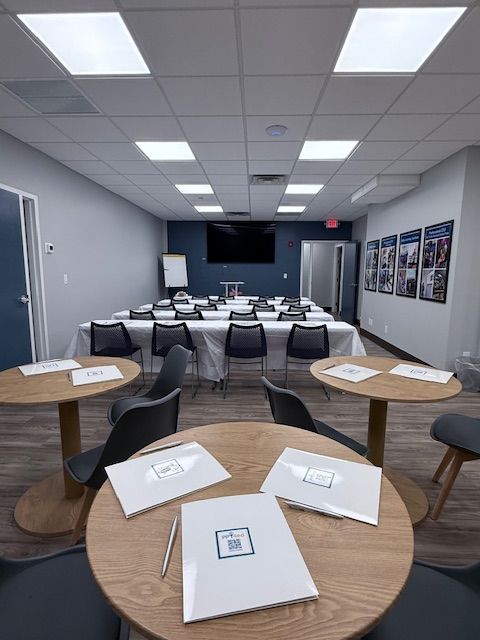 Meeting room with tables and chairs set up for a presentation. Several small tables with booklets are in the foreground.