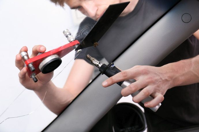 A person uses a red glass removal tool and wire to cut the adhesive sealant around a car windshield.