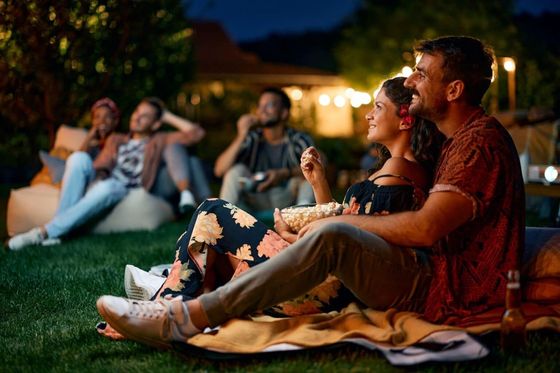 A group of people are sitting on the grass eating popcorn and watching a movie.