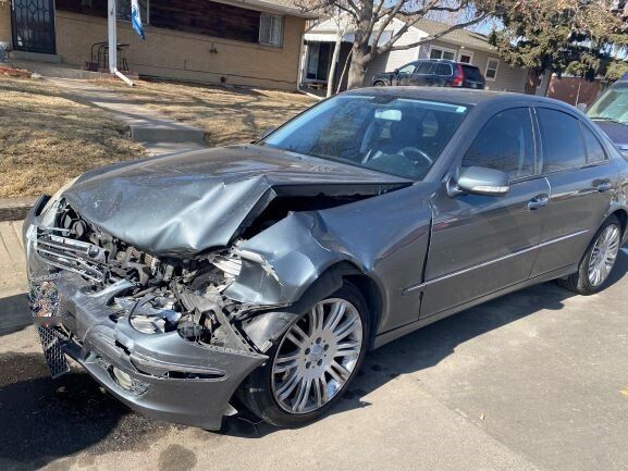 A gray car with a damaged front end is parked on the side of the road.