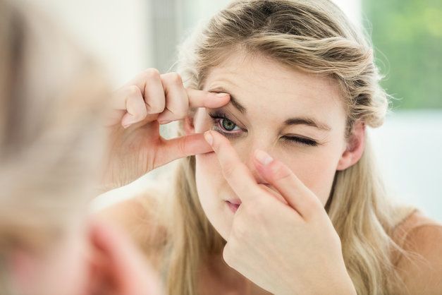 A woman is putting contact lenses in her eye in front of a mirror.