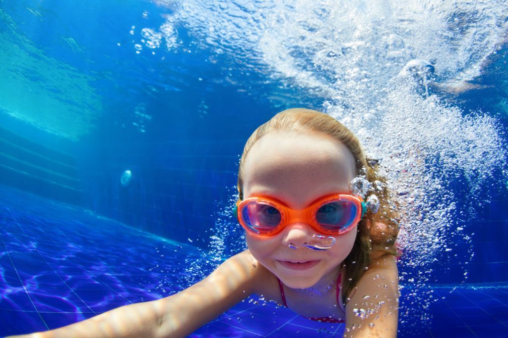 A little girl wearing goggles is swimming underwater in a pool.