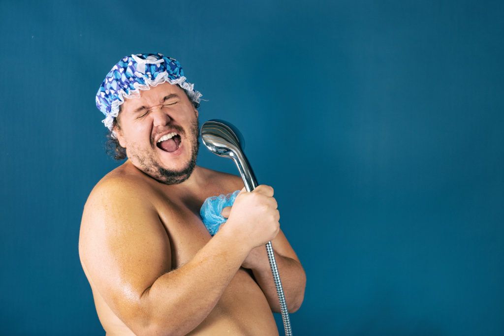 A shirtless man is singing into a shower head.