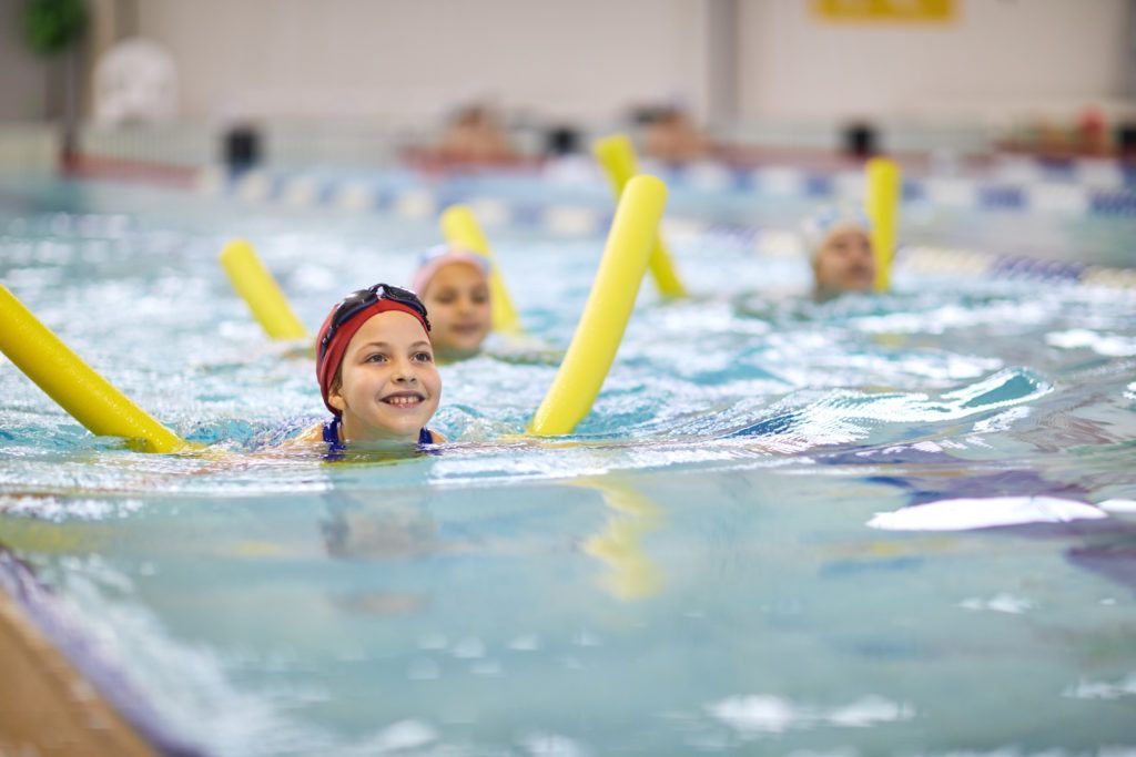 A group of children are swimming in a swimming pool with foam noodles.