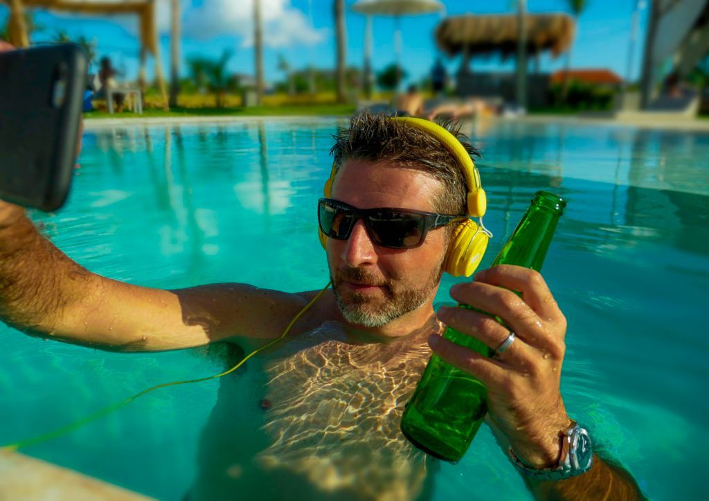 A man is taking a selfie in a swimming pool while holding a bottle of beer.