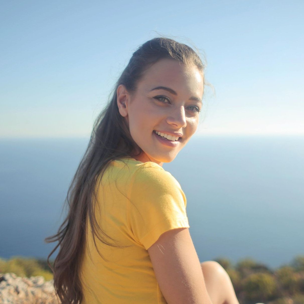 Woman in yellow shirt smiles over her shoulder at the camera; ocean and sky in background.