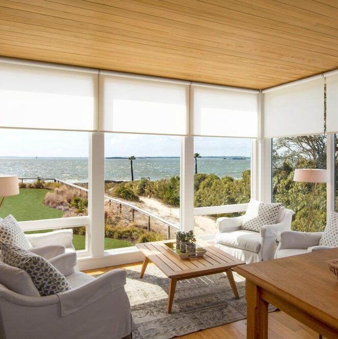 Sunroom with large windows overlooking a waterfront, featuring white seating, wooden table, and beige roller shades.