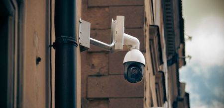 A security camera mounted on a building wall. The camera is white and black, with a rain gutter on the left.