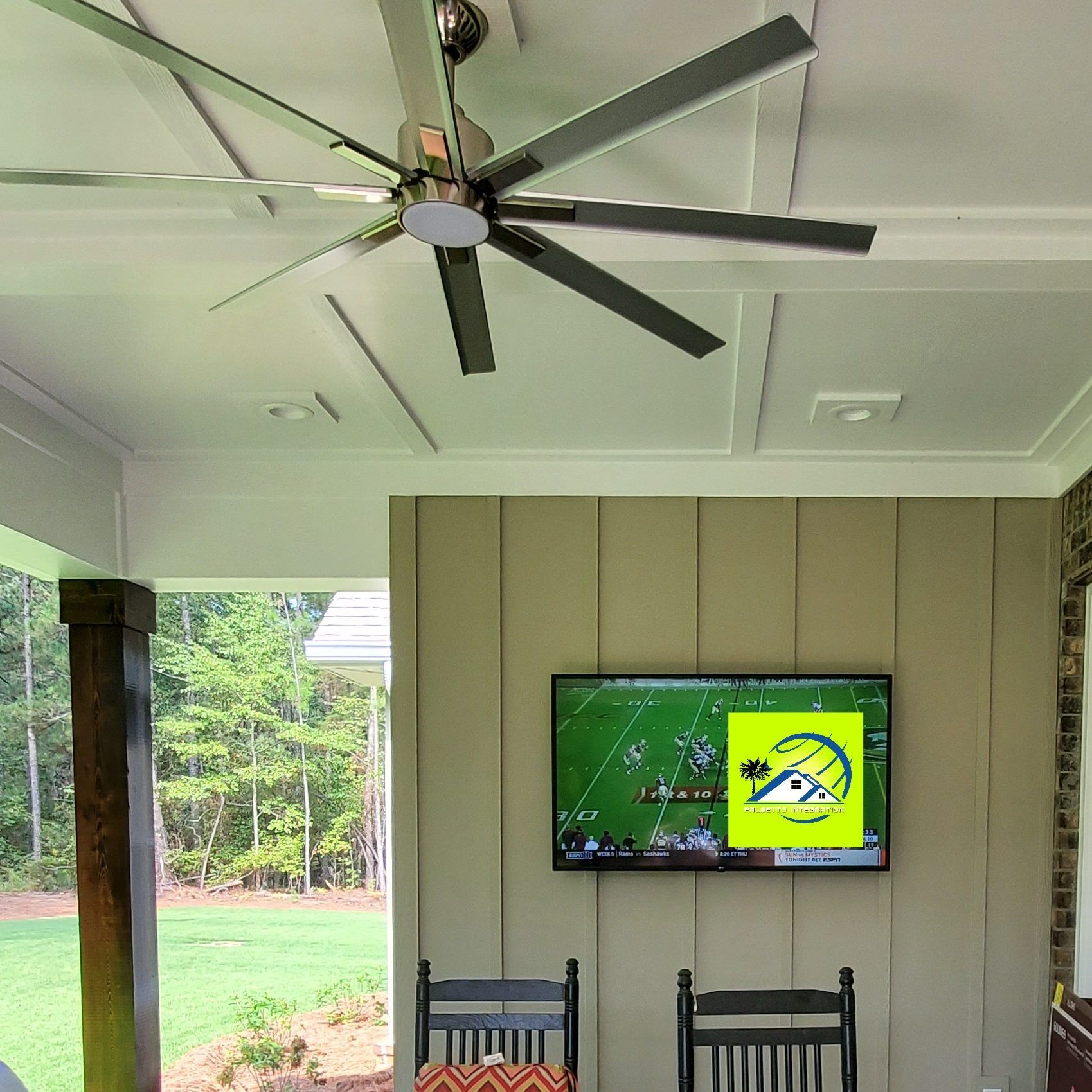 Outdoor space with a TV mounted on a light green wooden wall, two black chairs below, and a ceiling fan.