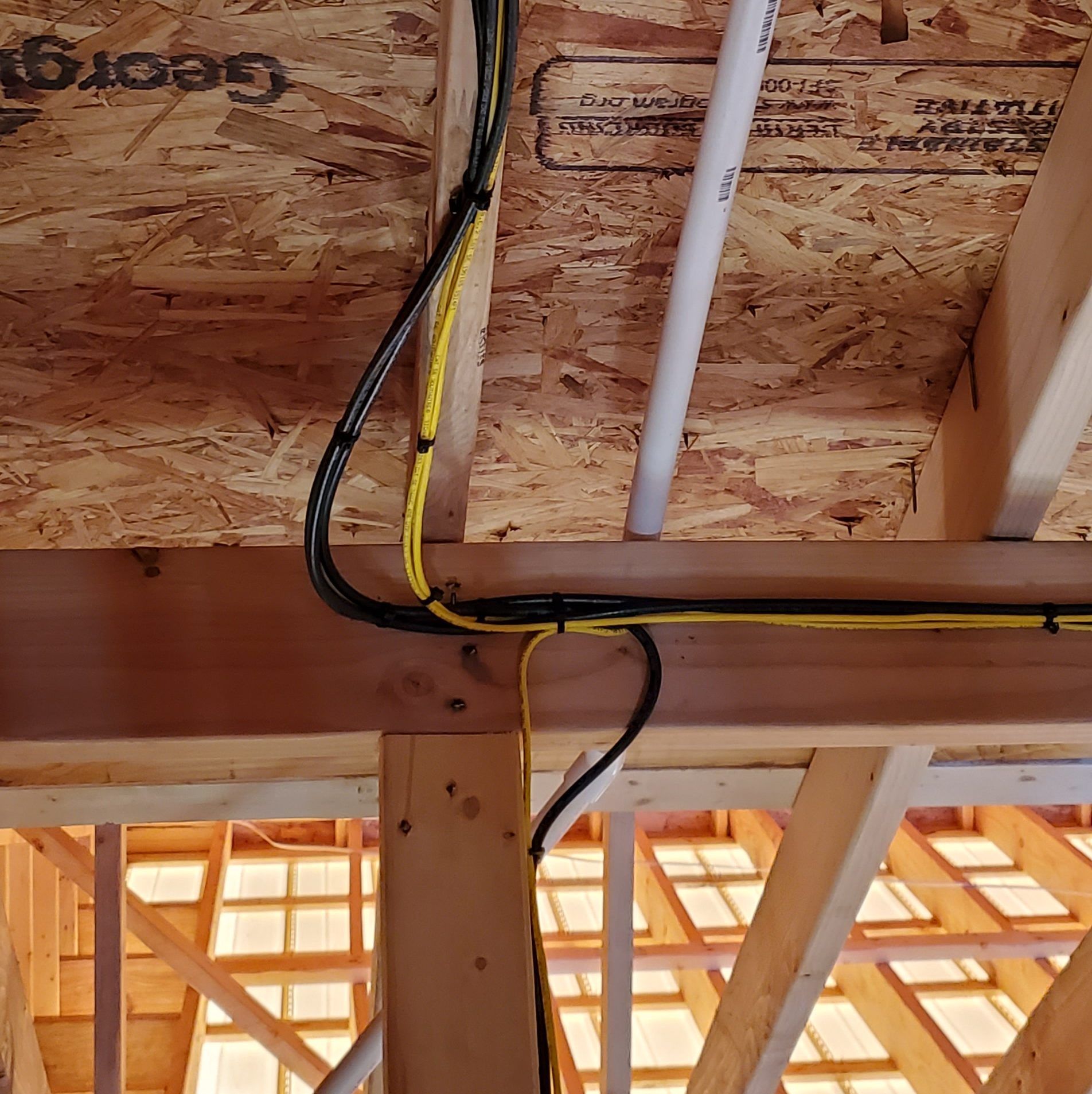Electrical wiring and a white pipe running along the wood ceiling and supporting beams in a building under construction.