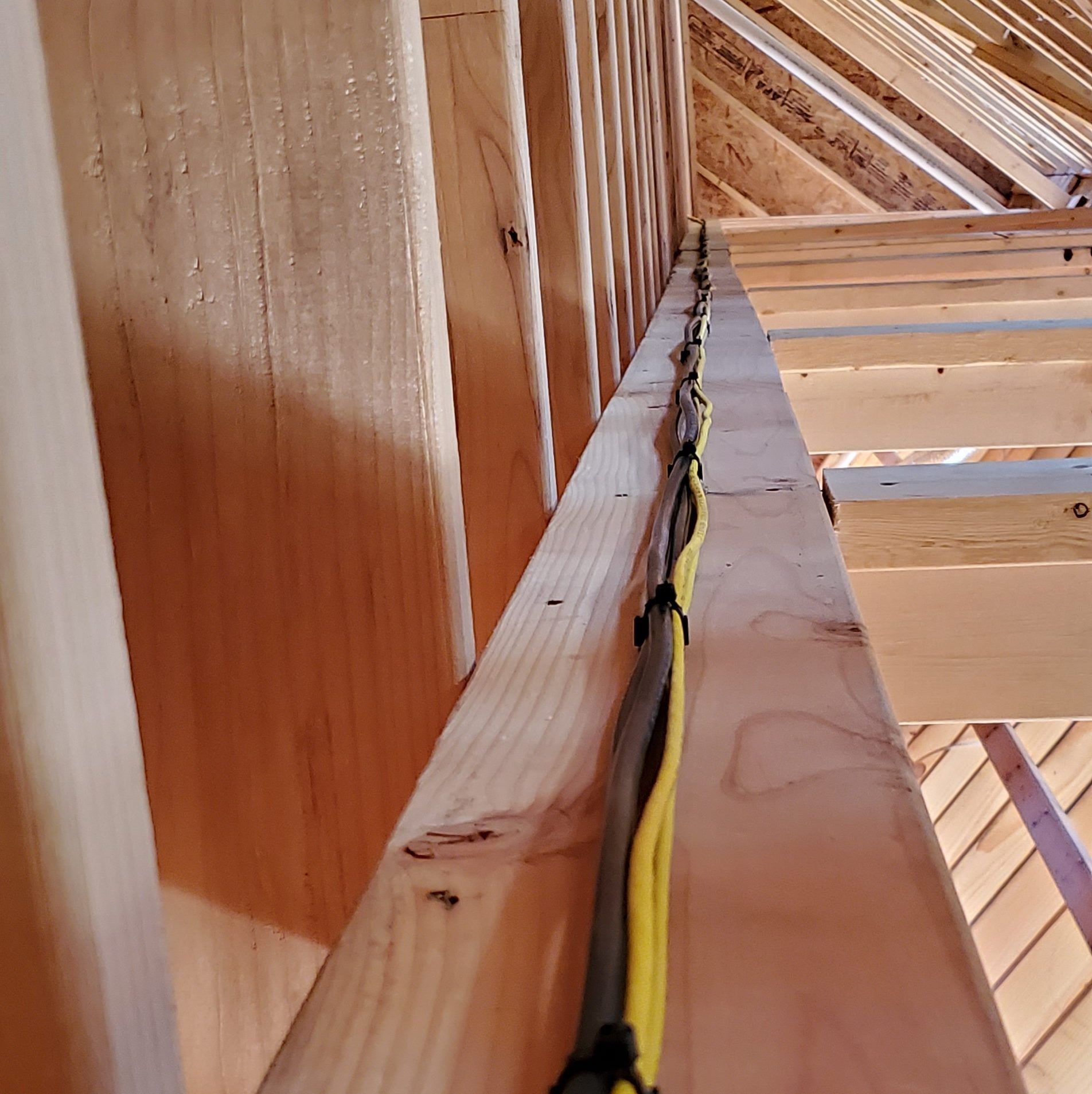 Electrical wires secured to a wooden beam within a building's framework. The wires are black, yellow, and grey.