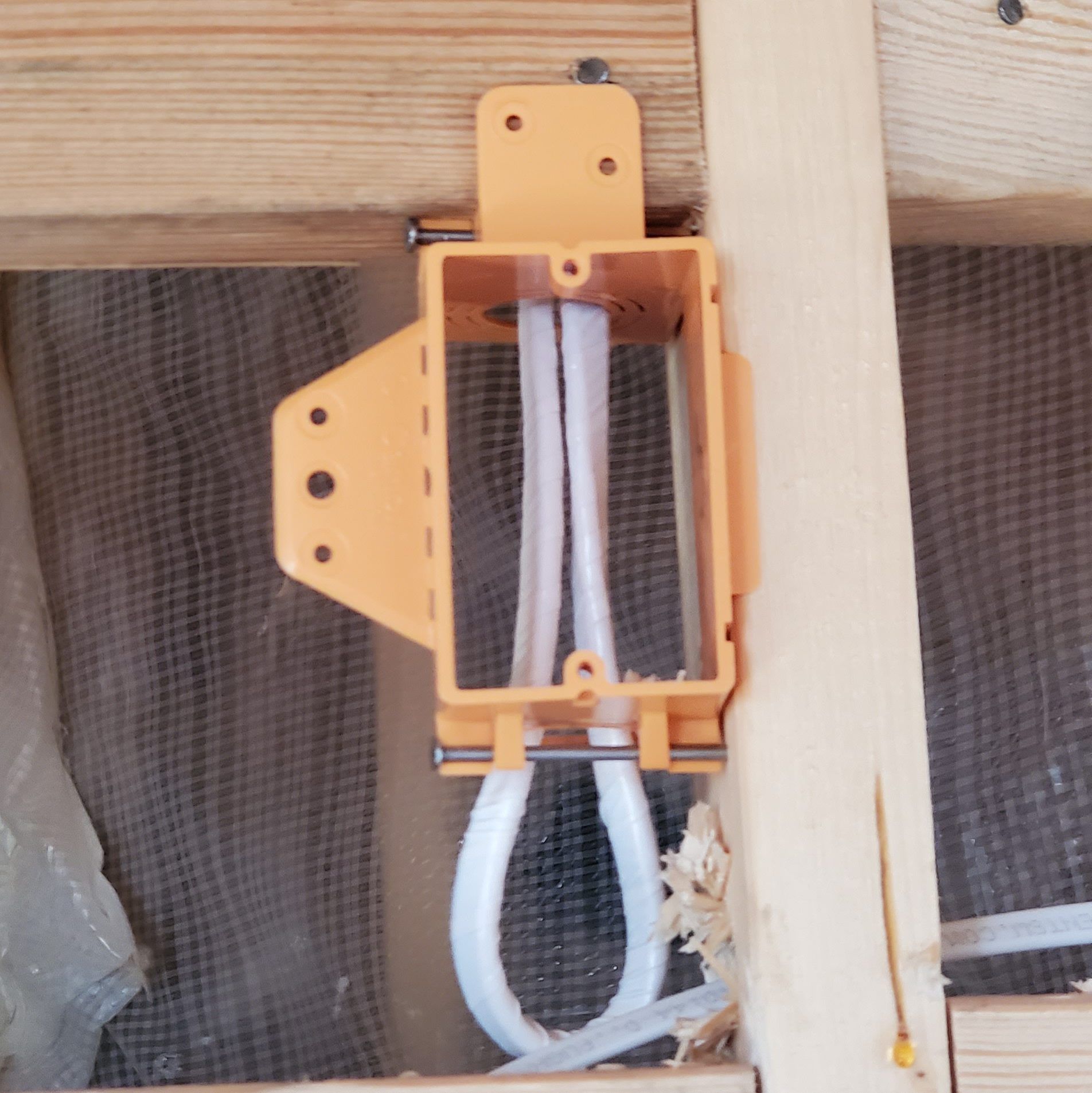 Orange electrical box mounted between wooden studs, with white electrical cables running through it.