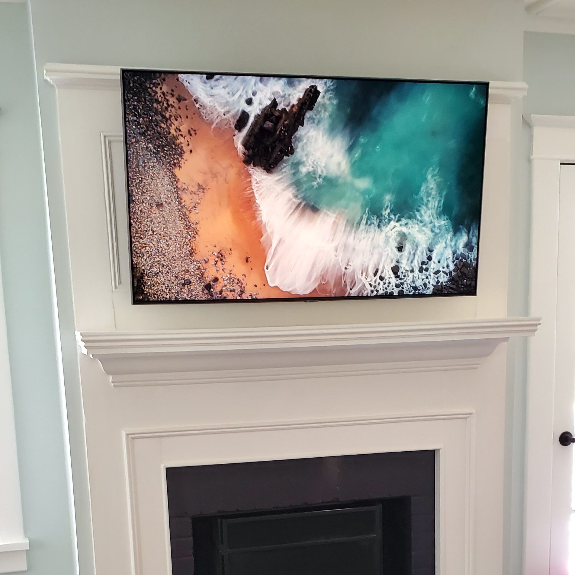 TV mounted above a fireplace, showing an ocean scene. The TV is surrounded by white molding against a light blue wall.