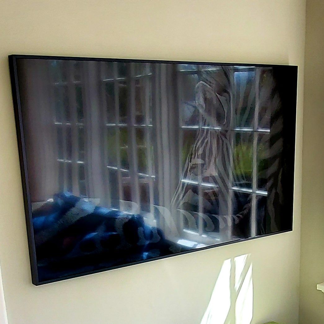 A wall-mounted black television reflecting a window with curtains and daylight.