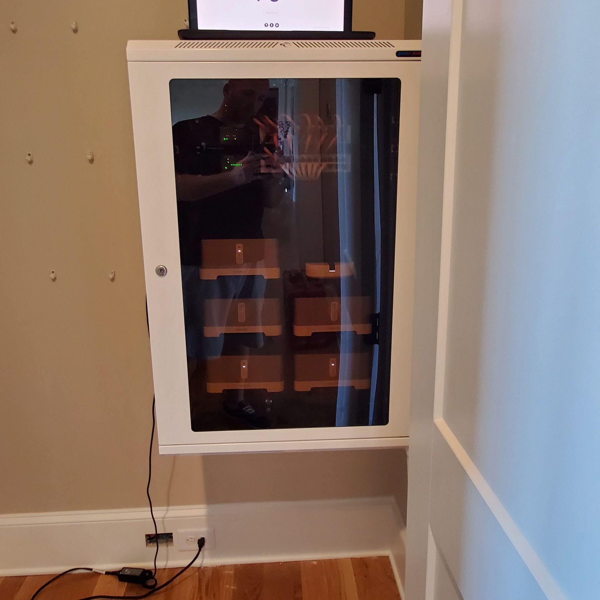 A white server rack mounted on a wall, with equipment visible inside a glass door. A laptop sits on top.