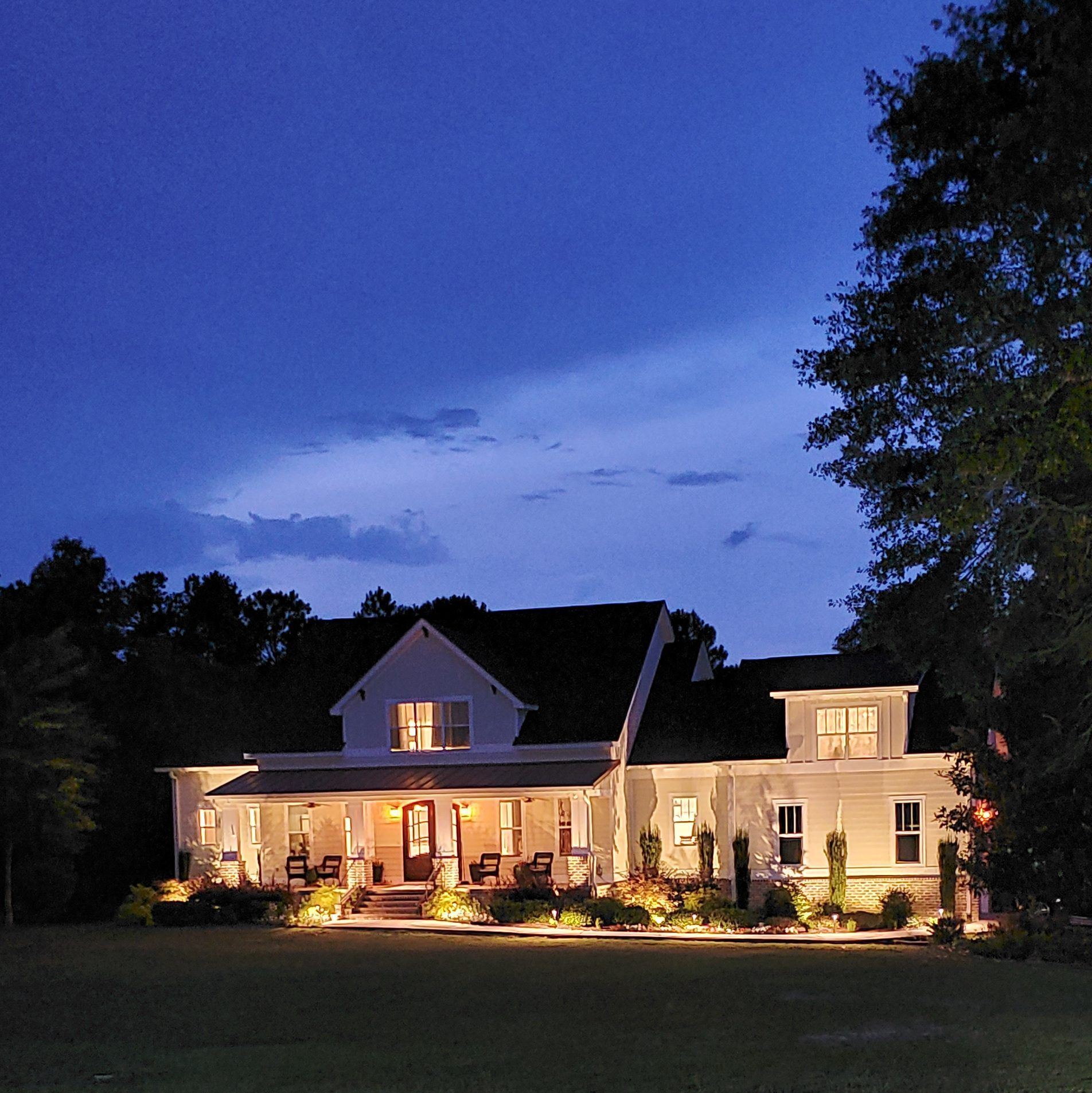 White farmhouse at dusk, illuminated by warm lights. Porch with rocking chairs and landscaping.