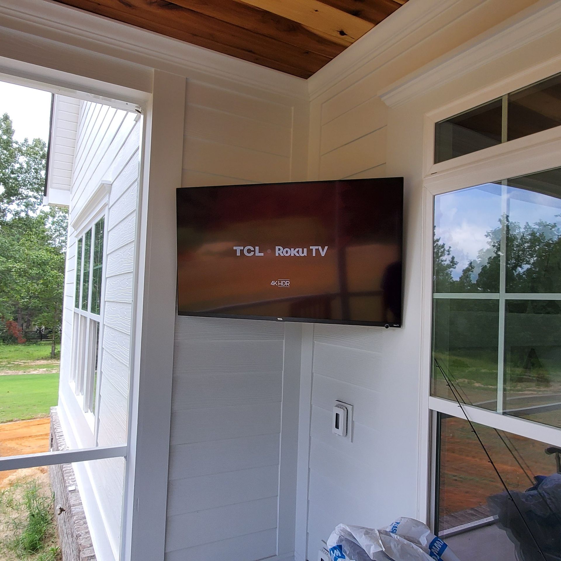 A mounted TCL Roku TV on a white paneled wall outside, next to a window and an electrical outlet.