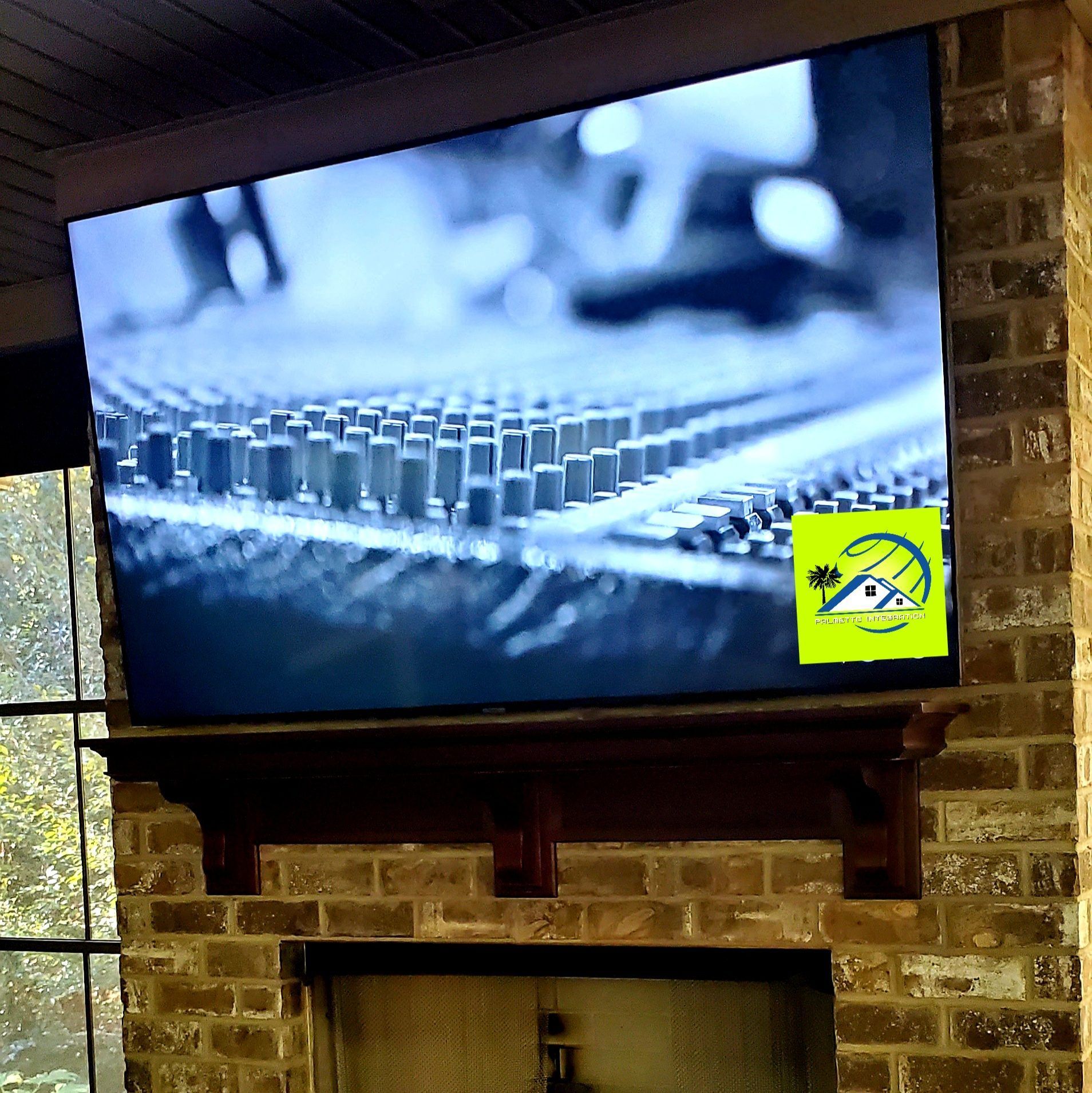 A large TV is mounted above a fireplace on a brick wall. The screen displays a close-up of circuit board components.