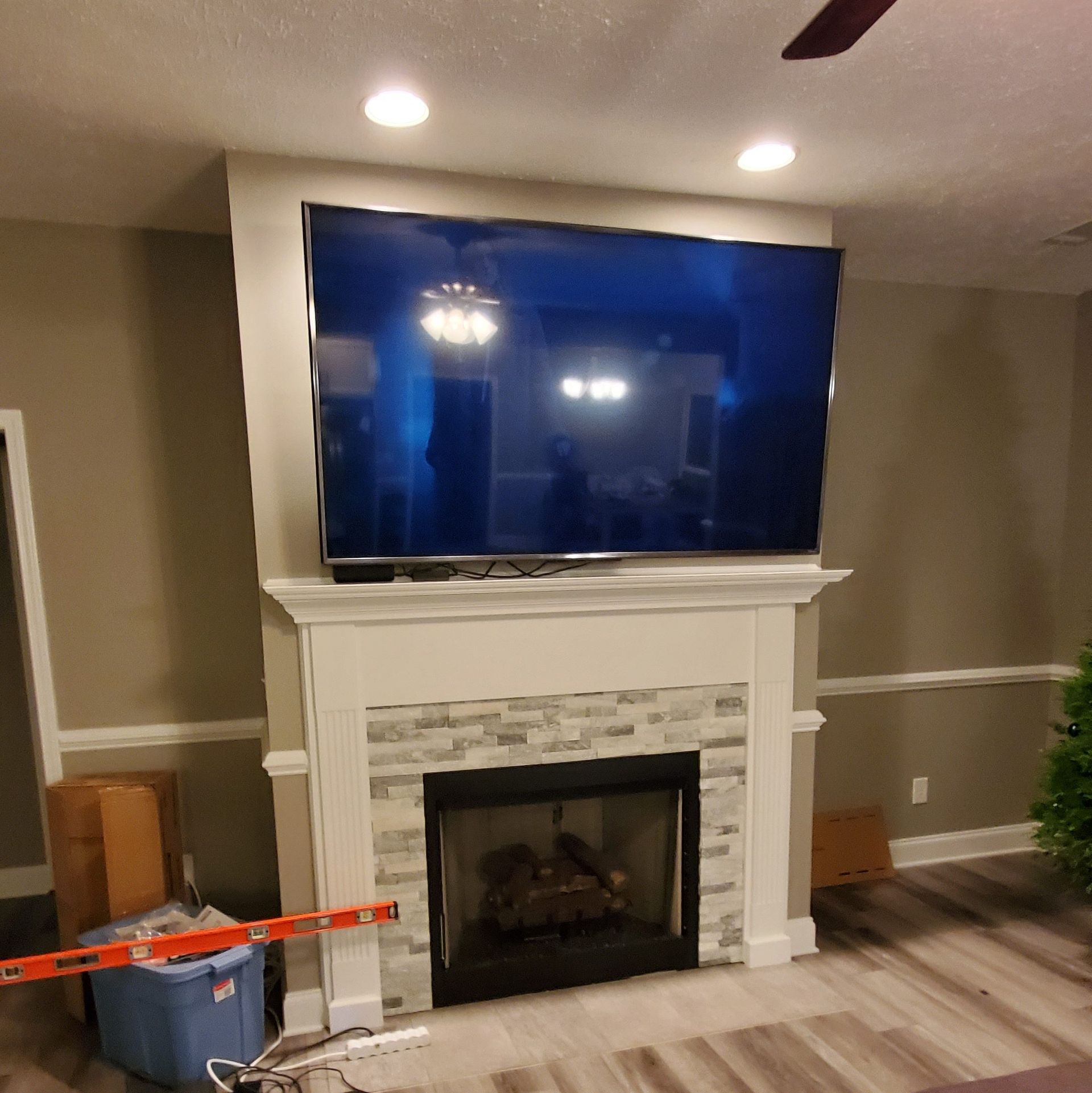 A large TV mounted above a fireplace with stone tile and white trim, in a room with neutral walls and recessed lighting.