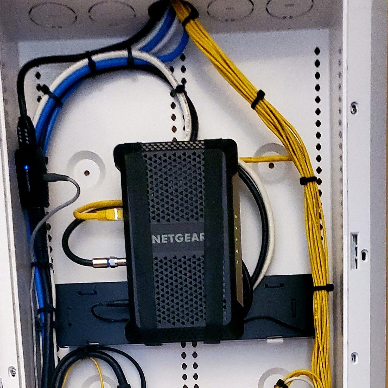 Network equipment, including a Netgear device, cabling, and power supply, installed inside a white wall-mounted enclosure.