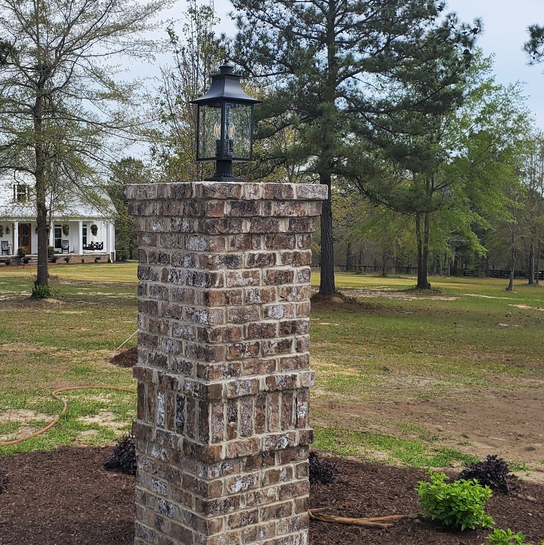 Brick pillar with a black lantern, in front of a white house and a grassy field.