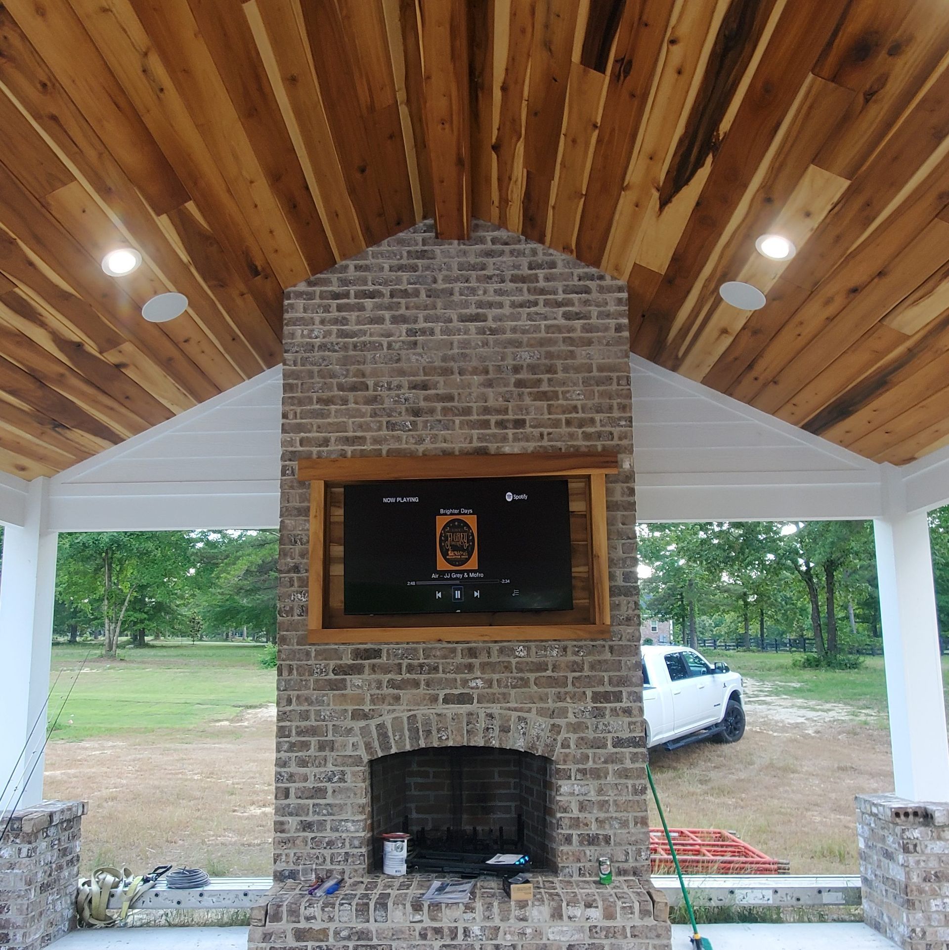 Outdoor brick fireplace with a TV and wood-paneled ceiling, a white truck parked in the background.