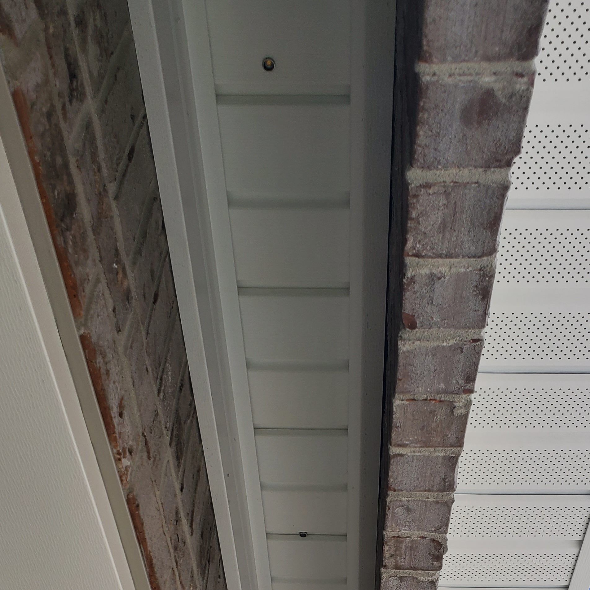Close-up view of house exterior showing white siding, brick, and perforated soffit. The siding has a nail in it.