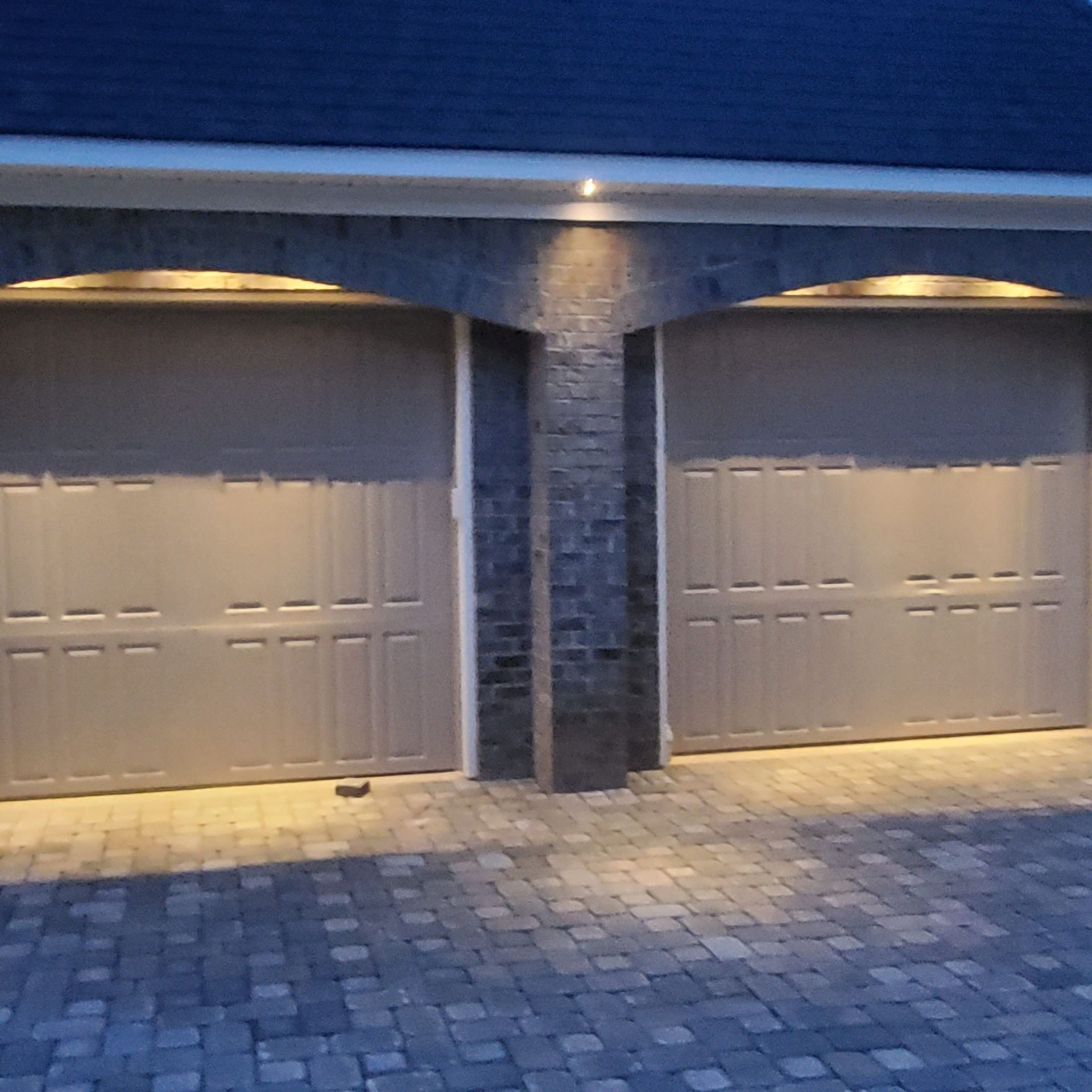 Two grey garage doors under brick arches, lit by spotlights, with a brick paver driveway.