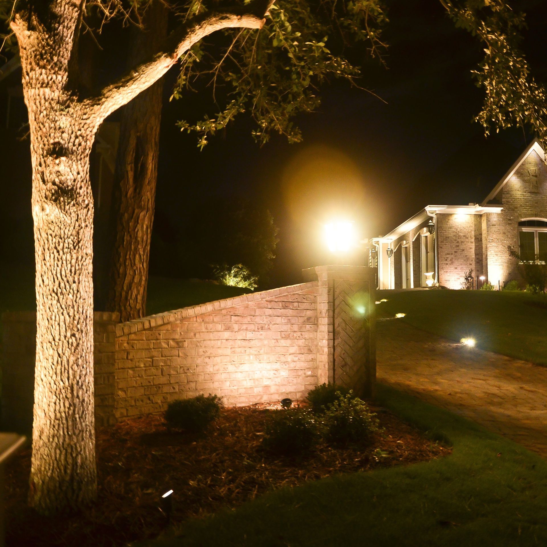 Nighttime shot of a home's illuminated landscaping. A large tree, brick wall, and house are lit with spotlights.