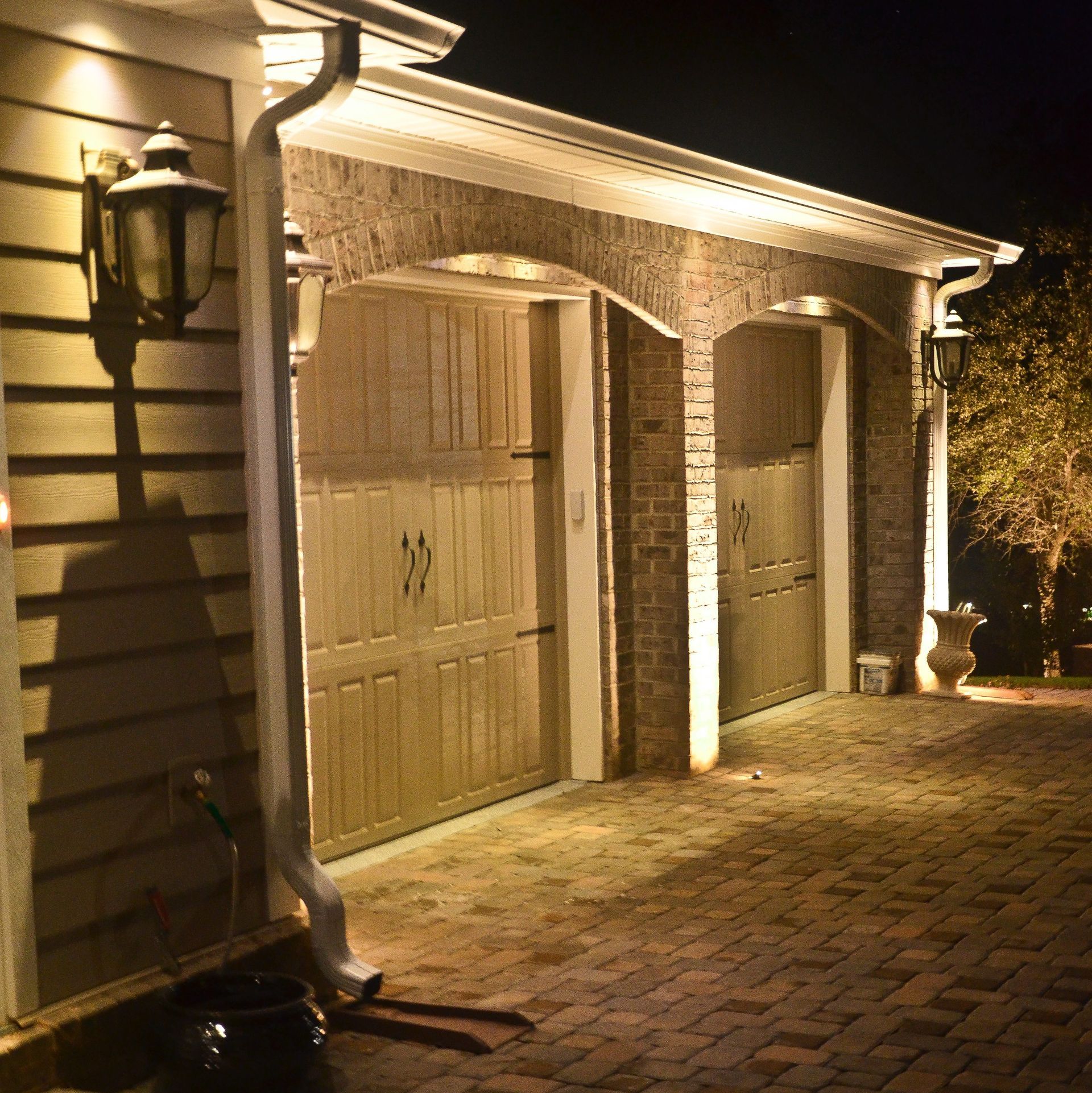Lit up garage with brick arches, wooden doors, and two lanterns at night, on a brick paved driveway.