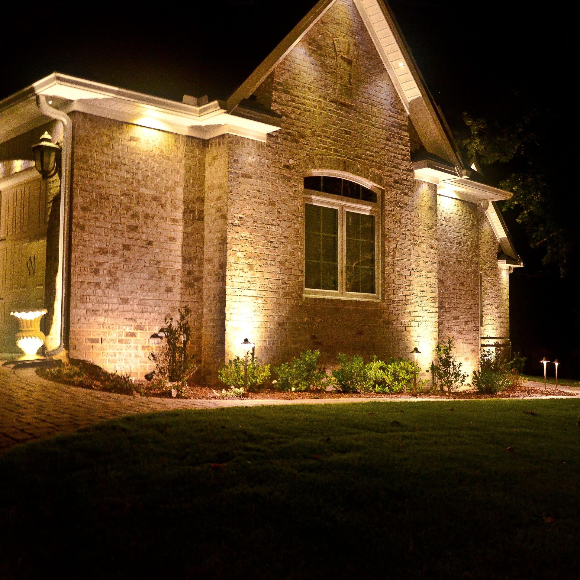 Brick house illuminated at night with spotlights, green lawn in foreground.