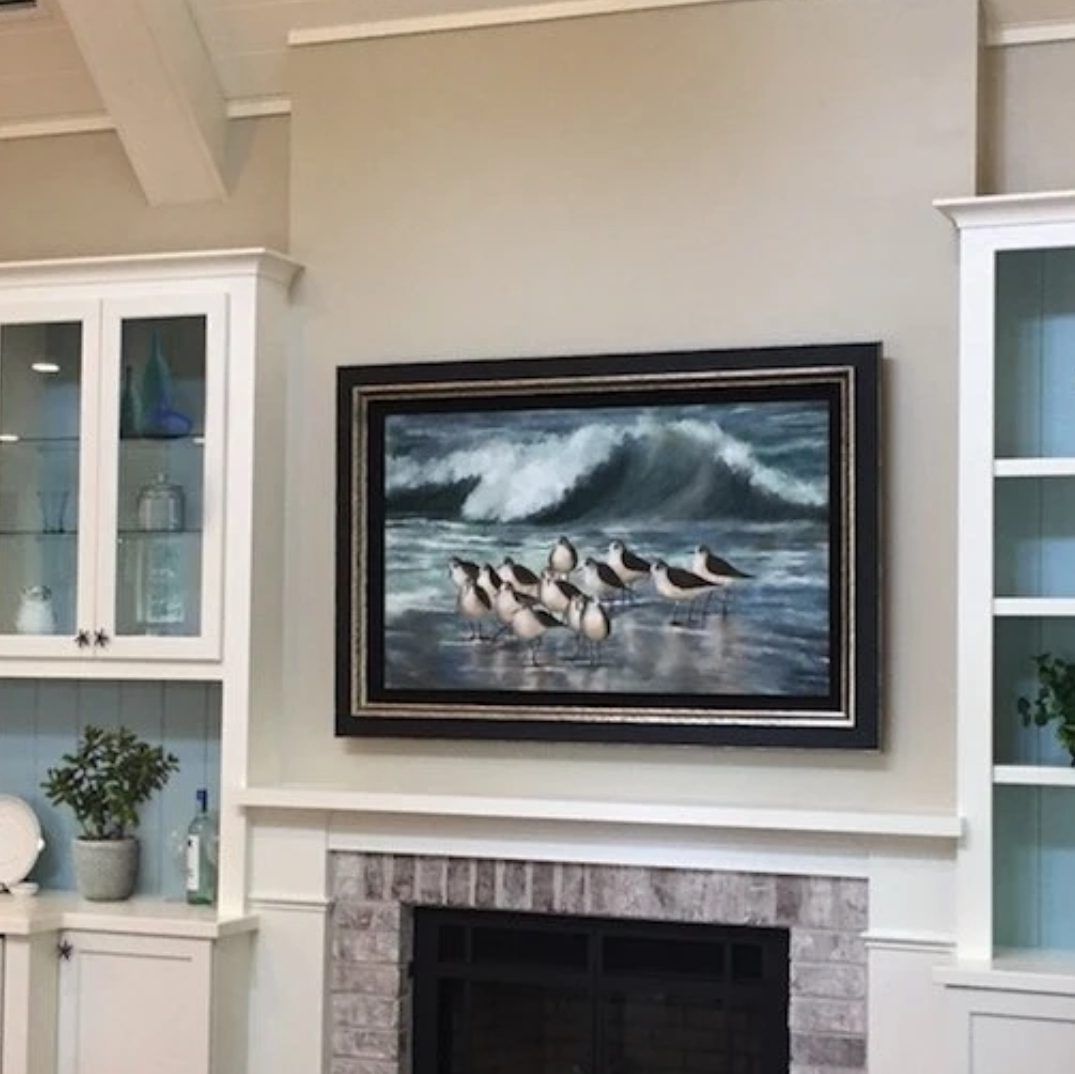 A framed painting of shorebirds on a beach with crashing waves above a fireplace, flanked by white cabinets.