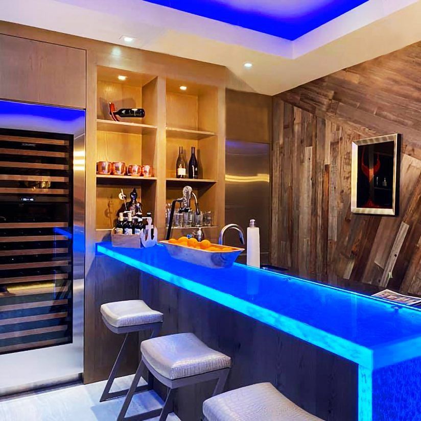 A stylish home bar with a blue-lit countertop, stools, and built-in shelving with drinks.